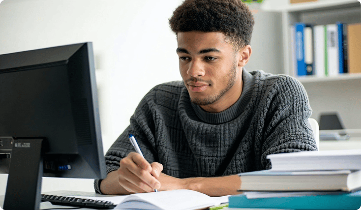 american young man take in notes while watching a videolesson on his computer his desk is clean and you can see his books and notes-1